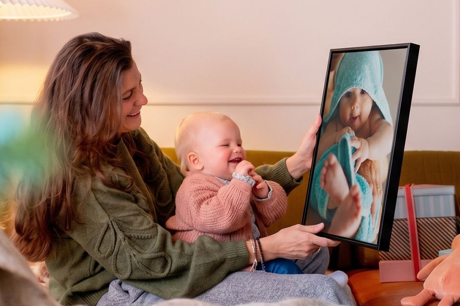 Mujer con un bebé en su regazo en un salón, sosteniendo una decoración de pared personalizada con una imagen del bebé mientras el bebé la mira y se ríe, para ilustrar las ideas de Hofmann para crear decoración de pared única con fotos del bebé en su primera Navidad.