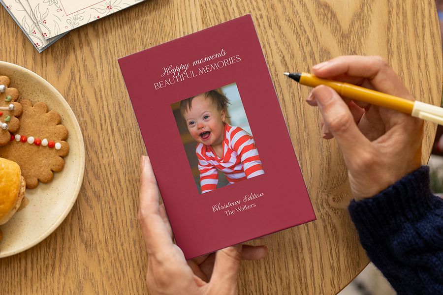 Manos a punto de firmar una tarjeta de Navidad personalizada con foto de un bebé sonriente, un mensaje navideño y el apellido de la familia en la portada, junto a un plato de galletas de Navidad sobre una mesa, para ilustrar las ideas de Hofmann para crear tarjetas de Navidad personalizadas para la primera Navidad del bebé.
