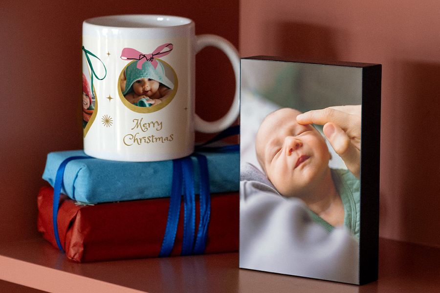 Taza personalizada con la foto de un bebé envuelto en una toalla y un mensaje de Feliz Navidad, sobre dos regalos envueltos junto a un bloque de fotos de un recién nacido dormido, para ilustrar las ideas de regalos con fotos de Hofmann para la primera Navidad de unos nuevos padres.