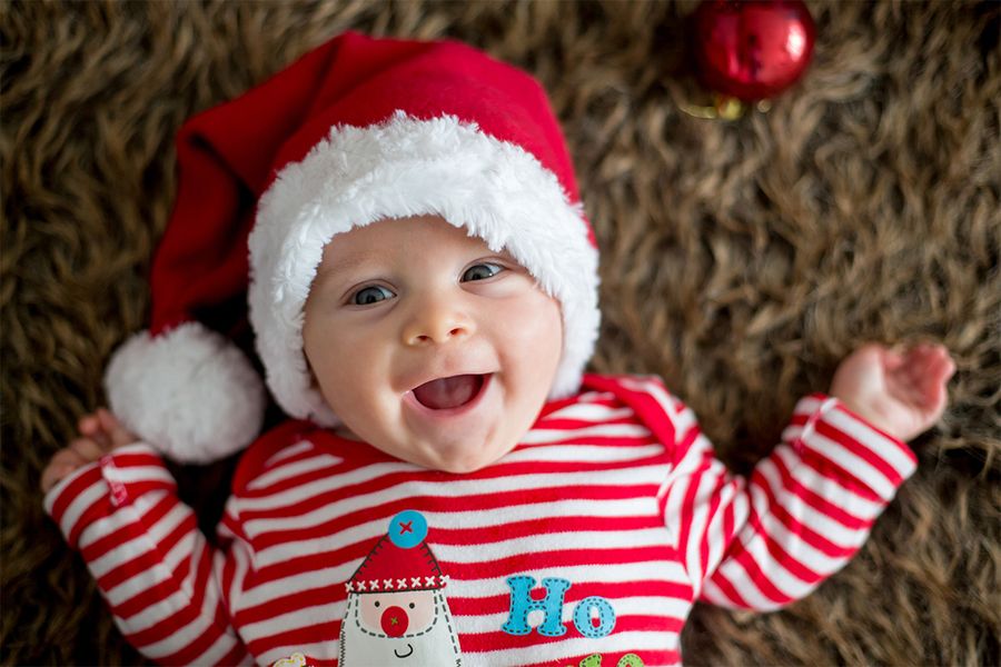 Bebé sonriente sobre una alfombra con un gorro de Papá Noel y una camiseta navideña de rayas rojas y blancas, para ilustrar los consejos de Hofmann para hacer fotos navideñas del bebé en su primera Navidad.