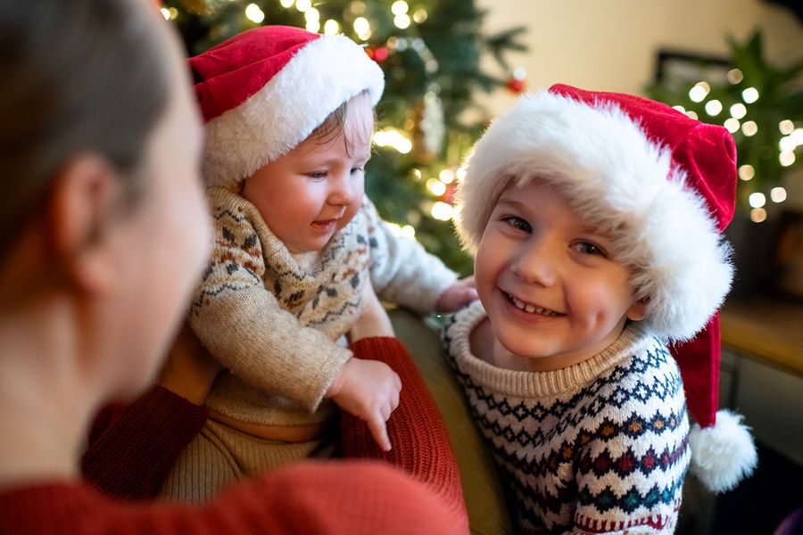 Mujer sosteniendo a un bebé junto a un niño sonriente al lado de un árbol de Navidad, con el bebé y el niño llevando gorros de Papá Noel y jerséis de invierno con estampado, para ilustrar los consejos de Hofmann para hacer fotos de Navidad inolvidables en familia y con el bebé.