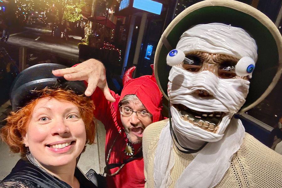 Tres adultos disfrazados de Halloween en la calle, uno con una máscara de esqueleto de ojos saltones, otro de diablo y una mujer con un sombrero negro, ilustrando las frases de Halloween de Hofmann para disfraces.