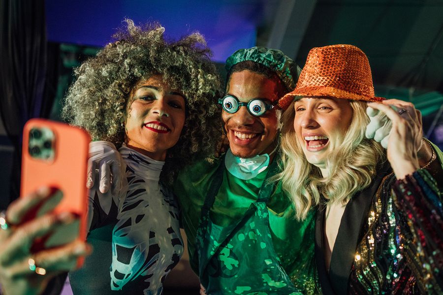 Tres adultos posan para un selfie en una fiesta de Halloween, una mujer con un disfraz de esqueleto, un hombre disfrazado de cirujano aterrador con unas gafas divertidas y una mujer con un sombrero de lentejuelas, ilustrando las frases de Halloween de Hofmann para grupos.