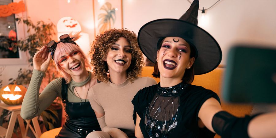 Tres mujeres disfrazadas en una fiesta de Halloween, una bruja, un animal y una cyberpunk, posan para un selfie con su móvil, presentando las 105 frases de Halloween para Instagram de Hofmann.