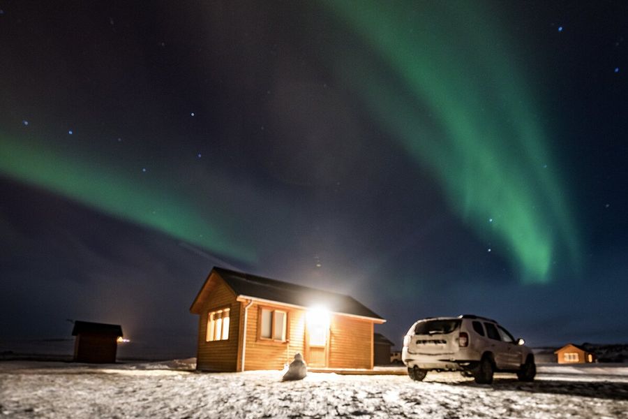 Una cabaña con una luz que brilla hacia fuera, junto a un coche, en un paisaje muy nevado, con auroras boreales verdes en el cielo por encima, ilustrando la explicación de Hofmann sobre qué son las auroras boreales y qué las provoca.