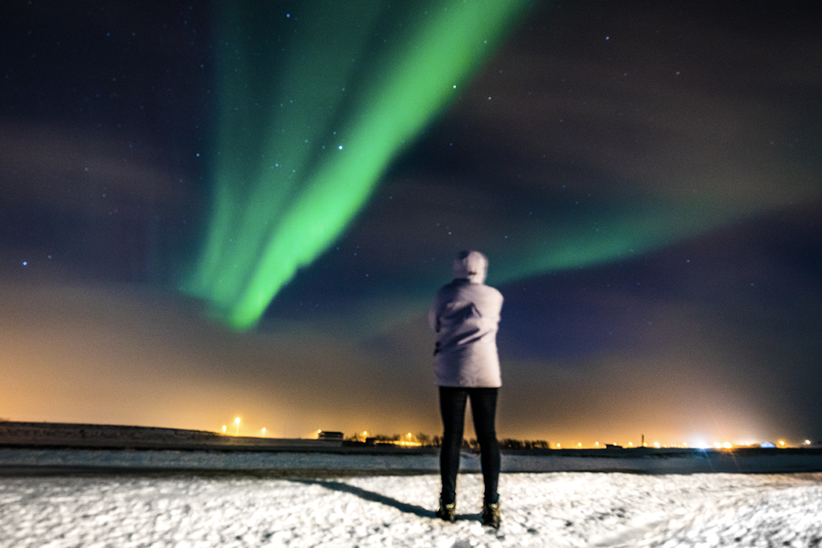 Una persona de pie en un paisaje nevado, mirando las auroras boreales verdes en el cielo, que ilustra los consejos de Hofmann para fotografiar la aurora boreal con el móvil.