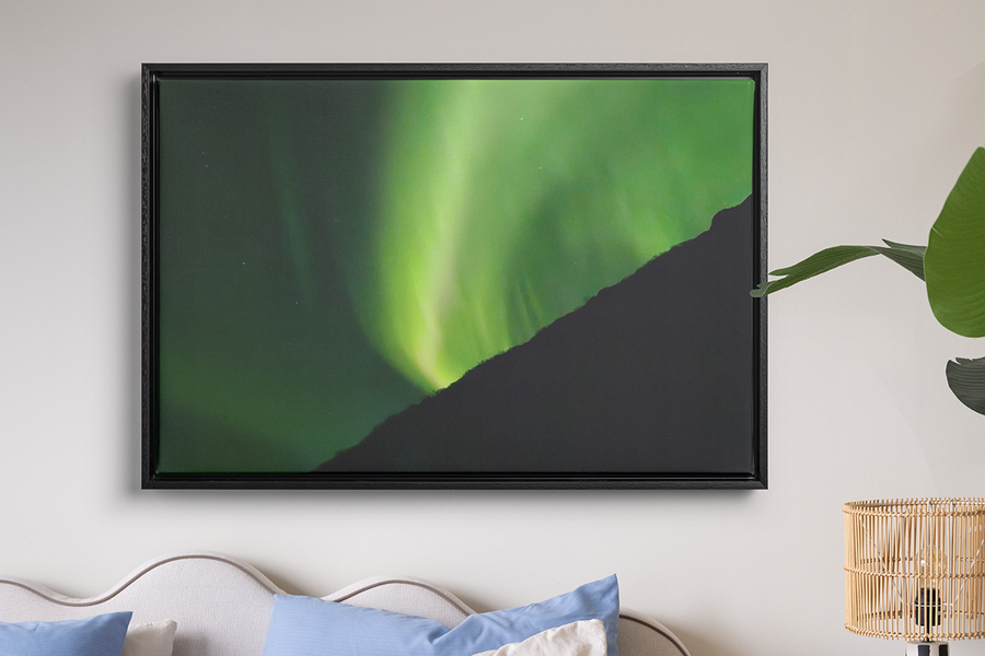 Una impresión enmarcada de auroras boreales muy verdes colgada en la pared sobre una cama y una lámpara de rafia, que ilustra la decoración de pared de Hofmann para convertir tus fotos de la aurora boreal en algo que merezca la pena conservar.