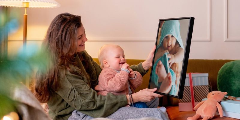 Mujer con un bebé en su regazo en un salón, sosteniendo una decoración de pared personalizada con una imagen del bebé mientras el bebé la mira y se ríe, para ilustrar las ideas de regalos con fotos de Hofmann para la primera Navidad del bebé.