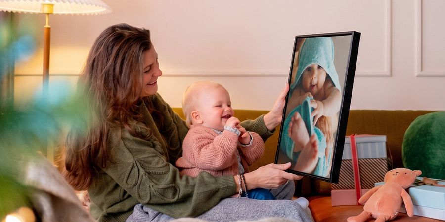 Mujer con un bebé en su regazo en un salón, sosteniendo una decoración de pared personalizada con una imagen del bebé mientras el bebé la mira y se ríe, para ilustrar las ideas de regalos con fotos de Hofmann para la primera Navidad del bebé.