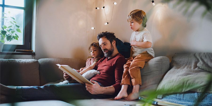 Un padre lee un libro a un niño y una niña pequeños en el sofá, con cálidas luces de hadas delante de ellos, presentando los consejos de Hofmann para una decoración otoñal acogedora al estilo hygge.