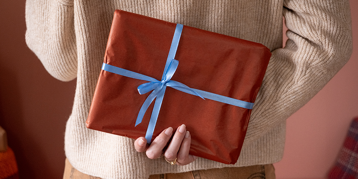 Mujer sosteniendo detrás de la espalda un regalo envuelto en papel marrón con un lazo azul, para ilustrar la guía de Hofmann con ideas de regalos de Navidad personalizados con fotos.