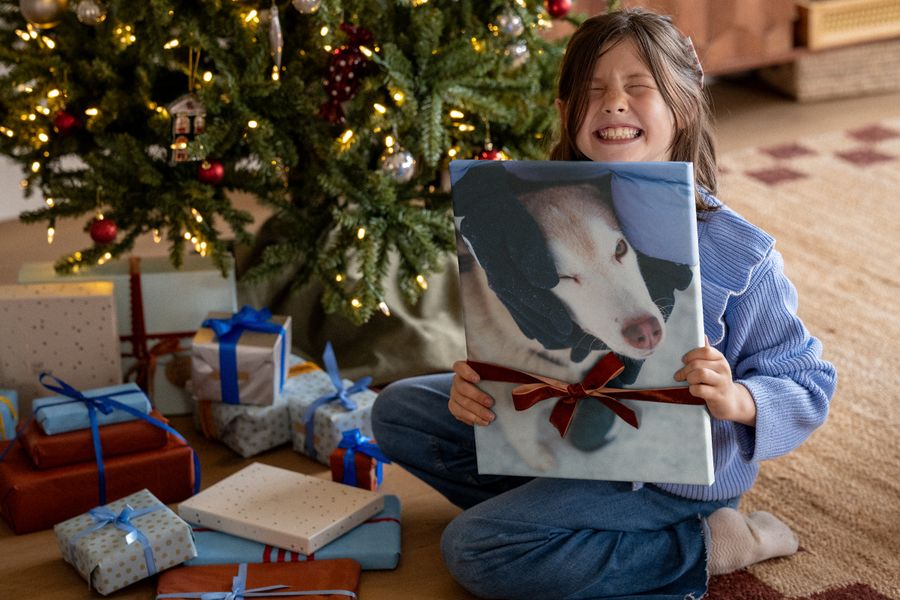 Una niña con jersey azul ríe a carcajadas mientras sostiene un lienzo con la foto de un perro, atado con una cinta roja, delante de un árbol navideño, ilustrando las ideas de decoración de pared de Hofmann para regalos de Navidad.