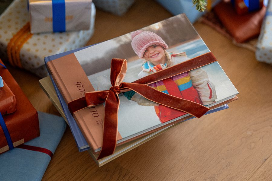 Pila de álbumes de fotos personalizados atados con un lazo de terciopelo marrón, con otros regalos envueltos al fondo, para ilustrar los consejos de Hofmann para preparar un regalo de Navidad personalizado con fotos.