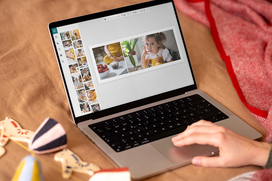 Una mano sobre el touchpad de un portátil diseña un álbum de fotos en pantalla, con una doble página de una niña y un bol de fresas, ilustrando el editor en línea de Hofmann para preparar regalos de Navidad en el Black Friday.