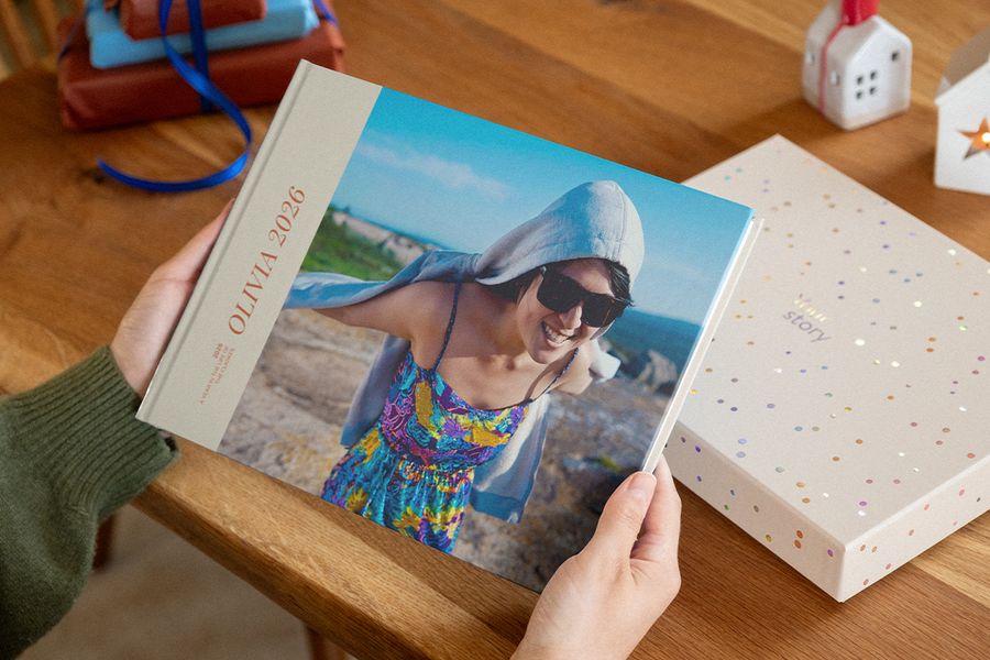 Dos manos sostienen un álbum de fotos con 2026 en la portada, en la que aparece una mujer sonriente con gafas de sol sobre una costa rocosa, junto a una caja de presentación con motivos de confeti, ilustrando los álbumes personalizados de Hofmann.