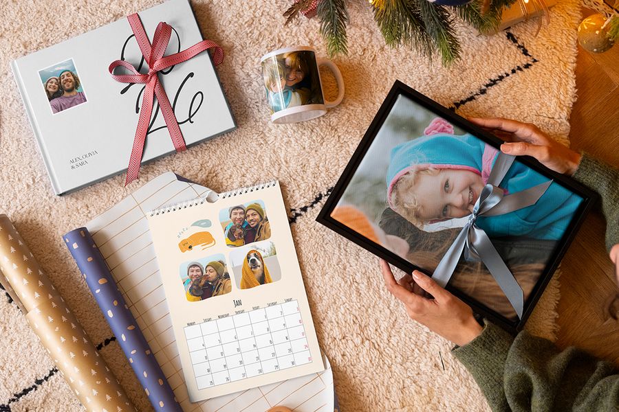 Selección de regalos personalizados de Hofmann, con decoración de pared de una niña pequeña con un lazo azul alrededor, una taza con una madre y su hijo, un calendario de fotos personalizado y un álbum de fotos de 2026 con un lazo rojo alrededor, junto a papel de regalo y con ramas de árbol de Navidad al fondo, para ilustrar los consejos de Hofmann para capturar los recuerdos adecuados en un regalo de Navidad personalizado.