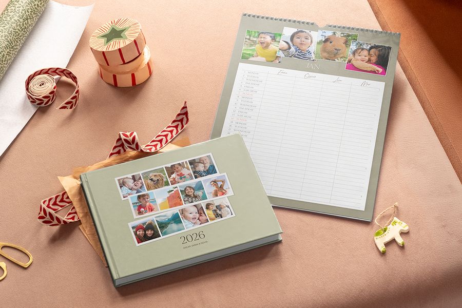 Un álbum de fotos verde salvia con 2026 en la portada está junto a un calendario familiar de pared con fotos arriba y columnas para cada miembro de la familia, ilustrando el calendario familiar compartido de Hofmann para regalos de Navidad en el Black Friday.