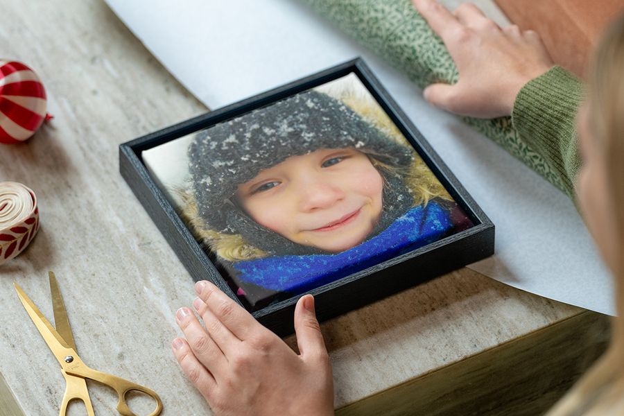 Lienzo enmarcado de una niña pequeña con ropa de invierno en la nieve, con las manos de una mujer listas para envolverlo, junto a papel de regalo, tijeras y lazo, para ilustrar los consejos de Hofmann para elegir un regalo personalizado con fotos que encaje con el estilo de cada persona.