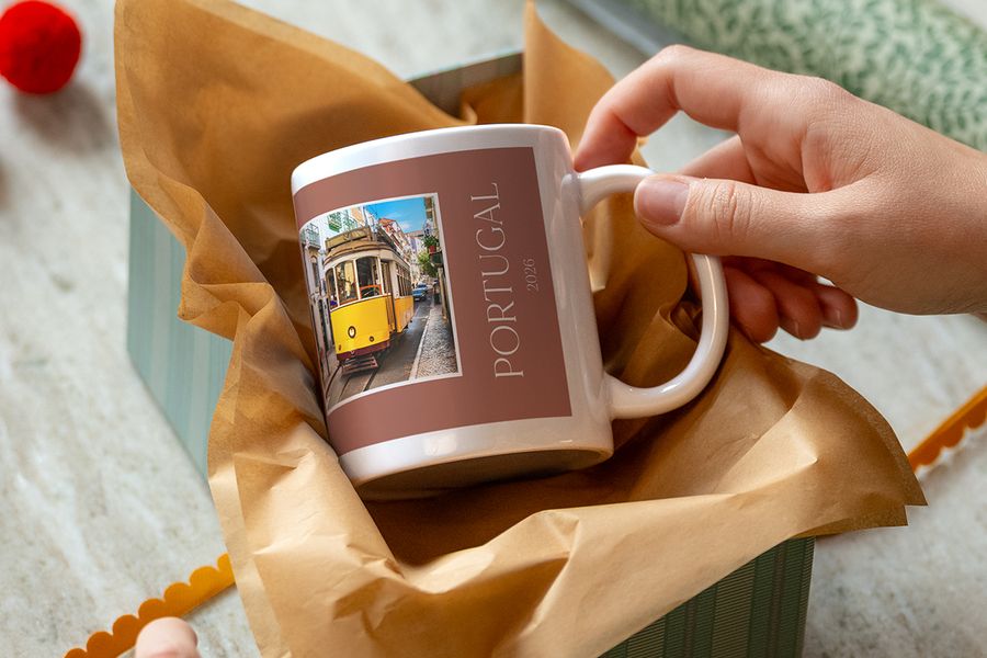 Una mano levanta una taza sobre papel kraft, estampada con un tranvía amarillo y el texto «Portugal 2026», ilustrando las tazas personalizadas de Hofmann como pista para un regalo de experiencia.