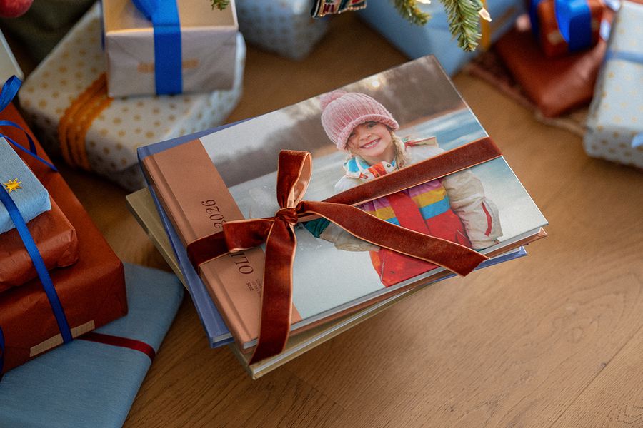 Una pila de álbumes de fotos atados con una cinta de terciopelo, y el de arriba muestra una niña sonriente con gorro rosa y el 2026 en el lomo, entre regalos de Navidad envueltos, ilustrando el álbum de fotos del año de Hofmann.