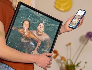 Una mujer sostiene su teléfono con el editor de Hofmann abierto mientras lleva bajo el brazo una decoración de pared de vacaciones con dos hombres divirtiéndose en una piscina, como inspiración para convertir fotos de vacaciones en un bonito cuadro de pared.