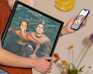 Una mujer sostiene un cuadro personalizado de Hofmann con una familia jugando en el agua durante las vacaciones, como inspiración para convertir fotos de vacaciones en una bonita decoración de pared.